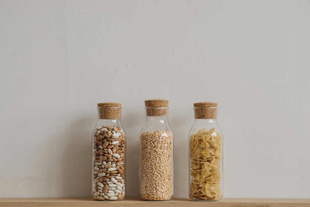 Three glass jars with cork lids holding beans, grains, and pasta on a wooden surface against a neutral backdrop.
